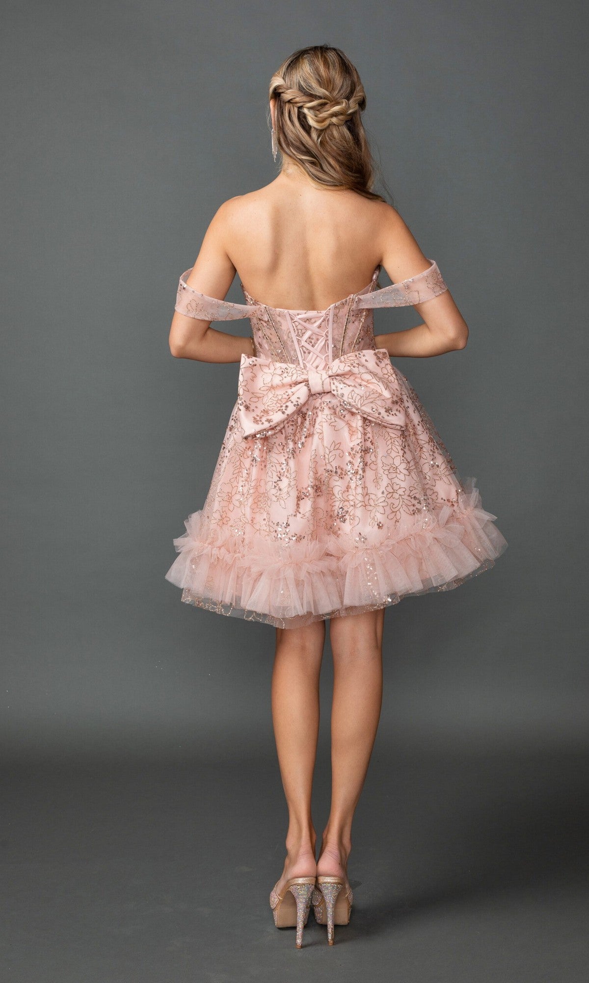 A woman faces away wearing the Dancing Queen 3422 short prom dress, featuring a strapless corset bodice, large bow, pale pink glitter details, and matching high heels.